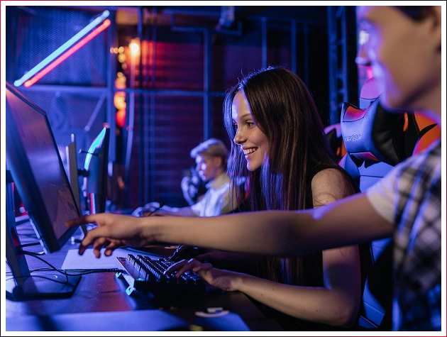 Usmívající se dívka hraje na počítači v herním klubu, zatímco chlapec v popředí ukazuje rukou na její monitor.